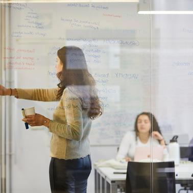A woman is presenting ideas on a glass board while others listen in a modern office environment.