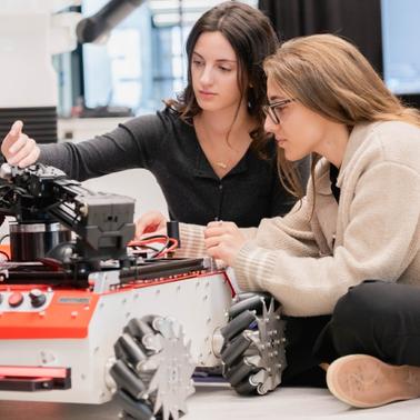 Two women are collaborating on a robotic project, focused on a red and white autonomous robot.