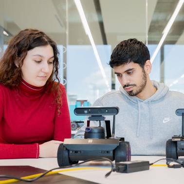 Two students are engaged in a discussion while examining robotic devices on a table.