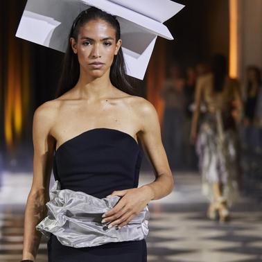 A model walks down the runway wearing a unique outfit with an oversized paper-like accessory on her head.