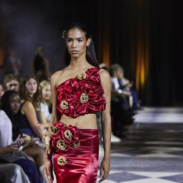 A model walks the runway wearing a striking red dress adorned with roses.