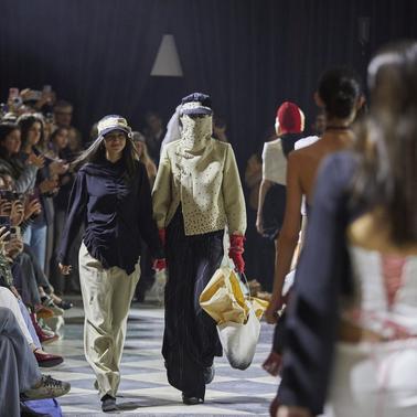 A fashion show featuring models walking down the runway with an audience in the background.