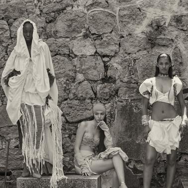 The image features three models posing in stylish attire against a textured stone wall.