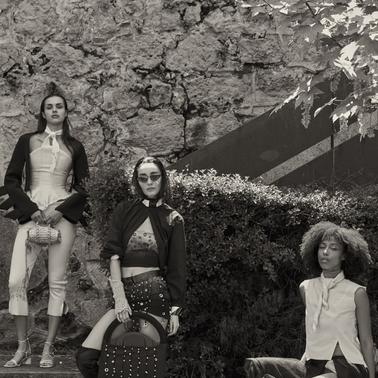 Three models showcase fashionable outfits against a textured stone wall.