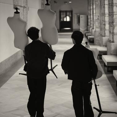 Two individuals are walking down a hallway carrying mannequin forms.
