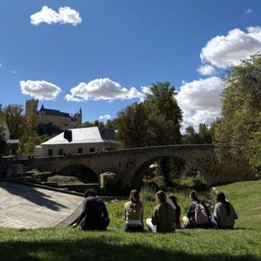 A group of people sitting on a grassy area near a stone bridge with trees and a castle in the background under a blue sky with clouds.