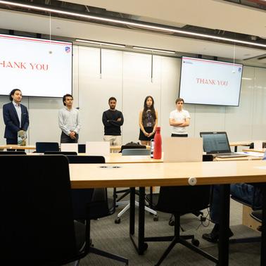 A group of five people stand in front of a screen that displays 'THANK YOU' while addressing an audience.