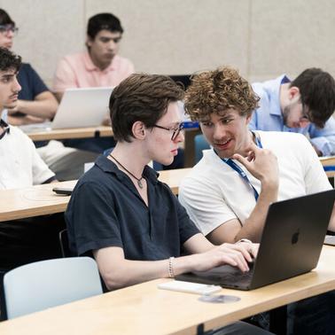 A group of students engaged in conversation while working on laptops in a classroom setting.