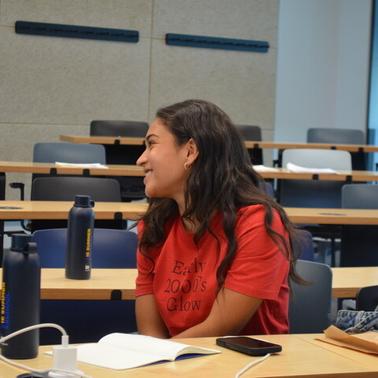 A smiling young woman wearing a red t-shirt is sitting in a classroom during a session.