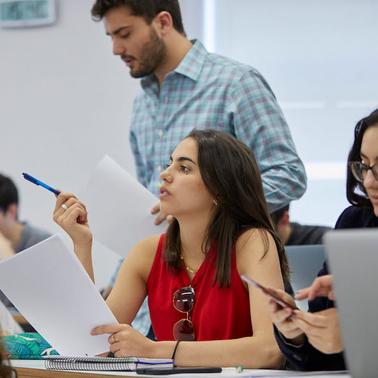 A group of students engaged in a classroom setting with one student speaking and others listening attentively.