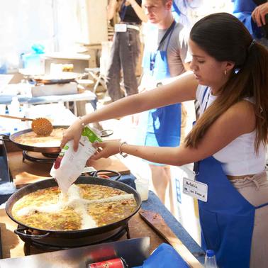 A woman in a blue apron is cooking in a large pan at a busy outdoor event.