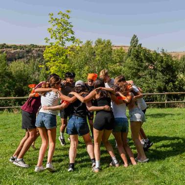 A group of people in a huddle outdoors in a grassy area with trees and a clear sky.