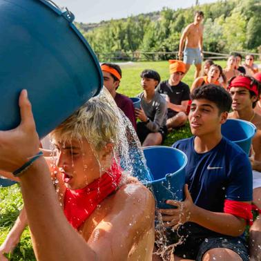 A young person is splashed with water from a blue bucket by another person in a sunny outdoor setting with a group of onlooking teenagers.