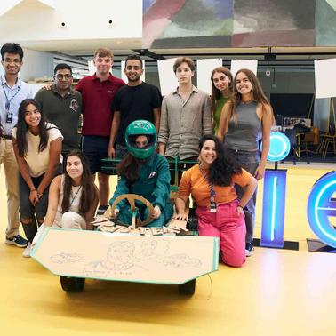 A group of diverse young adults gather around a table with a Google sign in the background at an apparent tech or educational event.