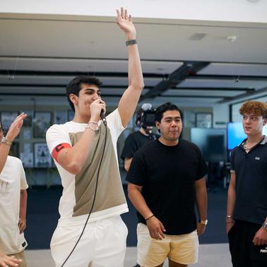 A young man enthusiastically singing into a microphone surrounded by cheering friends in a casual indoor setting.