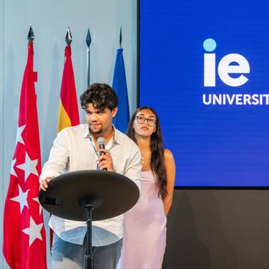 A young man is speaking at a podium with a microphone, while a young woman stands next to him at an event in a room with flags and a university's logo in the background.