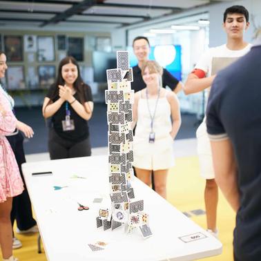 A group of people watching and smiling as a card tower is being built on a table.