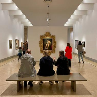 A group of people are viewed sitting on a bench in an art gallery, admiring the paintings displayed on the walls.