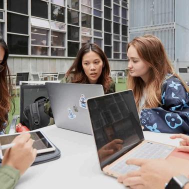 A group of young women studying together outdoors with laptops and tablets.