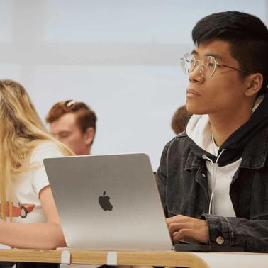 A young man with glasses and a denim jacket sits at a desk with a laptop in a classroom, looking to the side while other students are in the background.