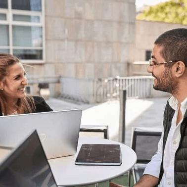 Two professionals smiling and engaging in a conversation at an outdoor table with a laptop and digital tablet.