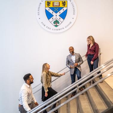 Four professionals are conversing on a staircase inside a building, beneath an emblem that includes the text 'Guildford College - For the Common Good'.