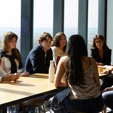 A group of seven people, including young adults and an older man, having a meeting around a table in a room with large windows.