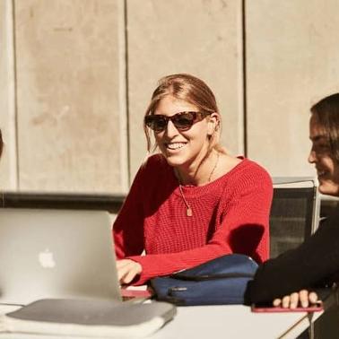 Three women sit at a table, smiling and talking with a laptop open in front of them.