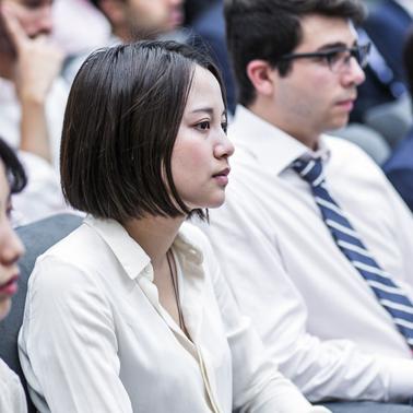 A group of young adults attentively listening in a seminar or class, predominantly wearing business attire.