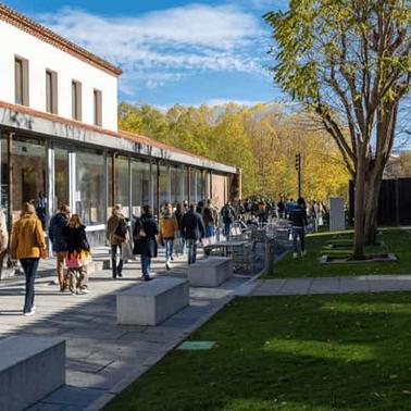 People walking near modern glass building in a park setting with trees and blue sky.