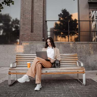 A woman sits on a bench outside a modern building, working on her laptop.
