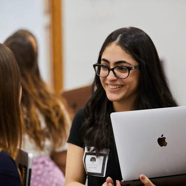A young woman with glasses is smiling and talking to her peers while holding a laptop at a gathering.