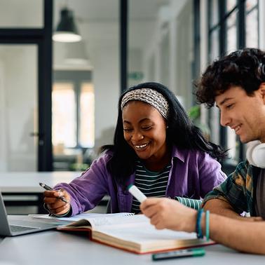 Two students are studying together at a table with a laptop and books.