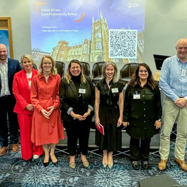 A group of nine individuals posing for a photo at an event, with a presentation screen displaying a QR code and the name 'School of Law, Queen's University Belfast' in the background.