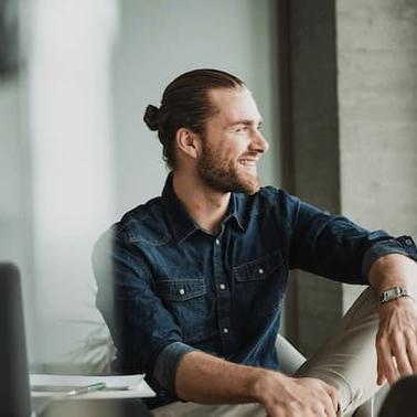 A man with a man bun smiling and looking out the window in a modern office setting.