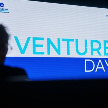 A person attends a Venture Day event at IE University, shown on a large presentation screen.