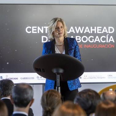 A woman delivers a speech at a podium with a backdrop displaying event logos and text.