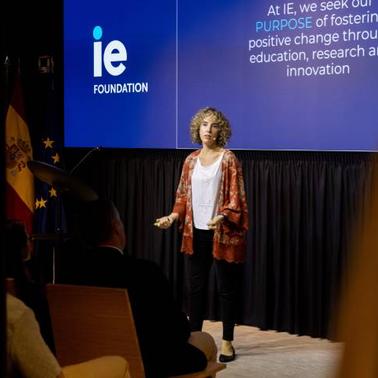 A woman presents at a conference in front of a banner that reads 'IE Foundation' with audience members in the foreground.