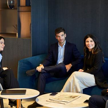 Four professionals in business attire are engaging in a conversation while sitting in a modern office lounge.