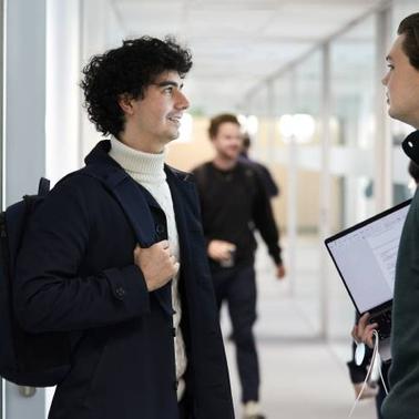 Two men talking in a corridor with one holding a backpack and the other holding documents.