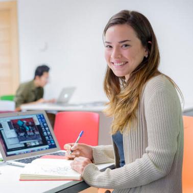 A young woman smiling at the camera while working on a laptop in a colorful study area.