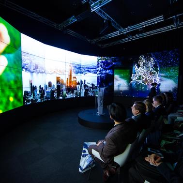 A large, modern presentation space with an immersive screen displaying various visuals and an audience seated in front.