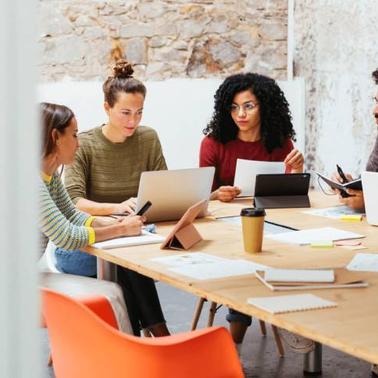 A group of four diverse professionals engaging in a discussion at a cluttered meeting table in a bright workspace.