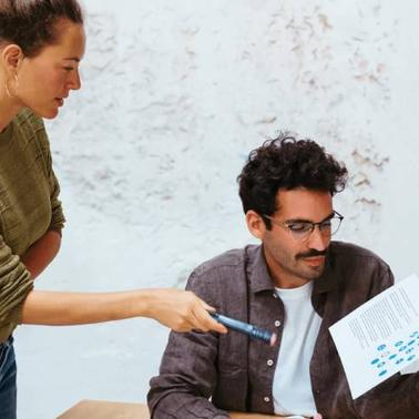 A woman pointing at a document held by a man sitting at a table in a casual office setting.