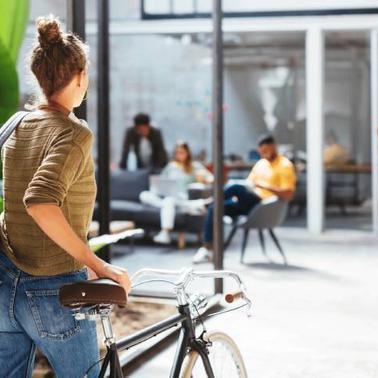 A person with a backpack standing beside a bicycle outside a modern office building, looking at people inside.