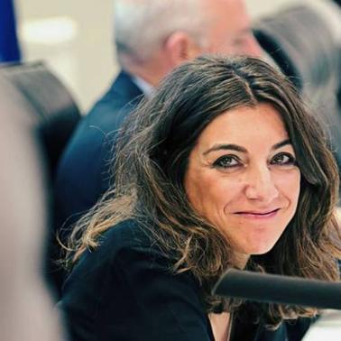 A woman in a professional setting smiles while sitting at a meeting with blurred figures in the background.