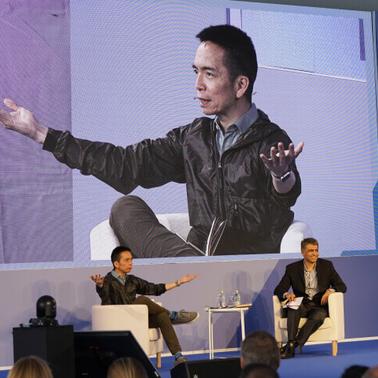 A man gestures while speaking at a conference, with a large screen behind showing his image, as another man listens seated beside him.