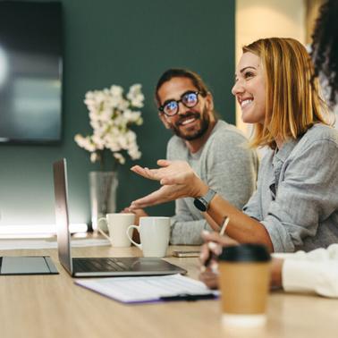 A group of diverse coworkers are engaged in a discussion around a table with laptops and coffee cups.