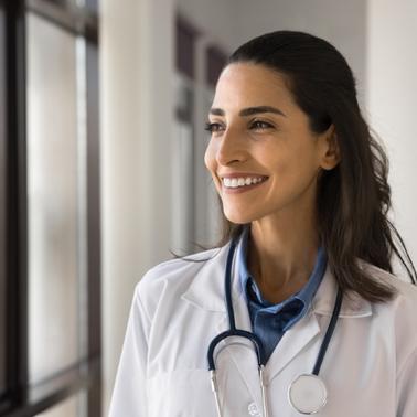 A smiling female doctor wearing a lab coat and stethoscope looks out a window.
