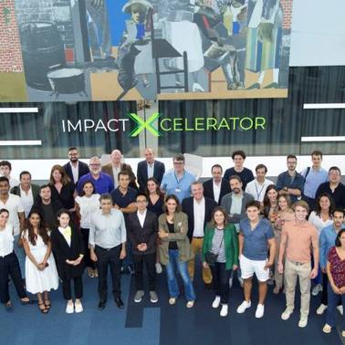 A large group of people posing for a photo in a modern building with a banner reading 'IMPACT XCELERATOR' in the background.